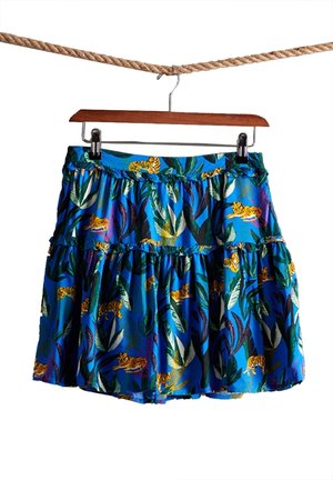 Jupe bleue avec imprimé tigre jaune et feuille verte suspendue à un cintre en bois accroché à une corde marron sur fond blanc.