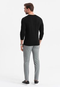 Uomo che indossa una maglietta nera a maniche lunghe, pantaloni grigi e sneakers nere, in piedi e con la schiena rivolta verso di noi, su uno sfondo bianco.