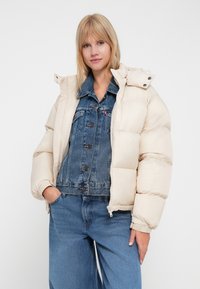 Puffjacka i ljusbeige med dragkedja och huva. Bärs över en denimjacka, ihop med vida blå jeans.