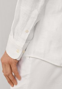 Mano izquierda al lado de la cadera con una camisa blanca de lino de manga larga con puño abotonado y pantalones blancos.