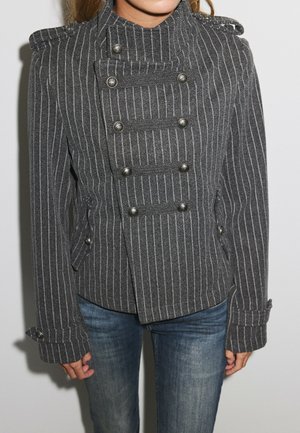 Donna che indossa una giacca militare grigia aderente a righe sottili con doppia fila di bottoni e jeans blu su sfondo semplice.