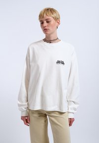 Weißes Langarm-T-Shirt mit Rundhalsausschnitt, mit einem schwarzen Grafikdruck auf der linken Brust. Getragen mit hellbeigen Hosen.