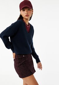 Granatowy sweter dzianinowy z dekoltem w kształcie litery V, zestawiony z bordową czapką oraz teksturowaną, wielokolorową spódnicą mini z tweedu z guzikami z przodu.