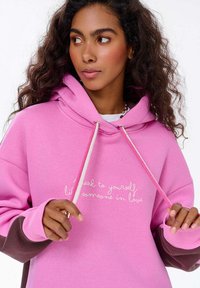 Růžová mikina s kapucí na šňůrku, se bílým vyšitým textem. Manžety v tmavší barvě, ukazující uvolněný design.
