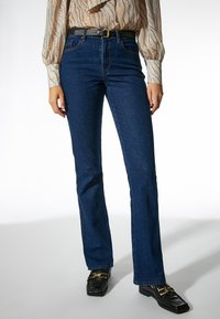 Jeans de mezclilla azul acampanados con una textura suave, cinco bolsillos y costuras en contraste, combinados con una blusa estampada y mocasines negros.