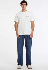 Młody mężczyzna stoi przodem, ubrany w białą koszulkę Guess Jeans, niebieskie dżinsy i białe trampki na jednolitym białym tle.