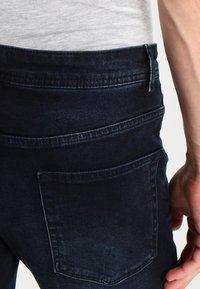 Tummansininen farkut, joissa on sileä pinta, taka-tasku ja hillityt tikkaukset. Suunniteltu keskitason vyötäröllä.