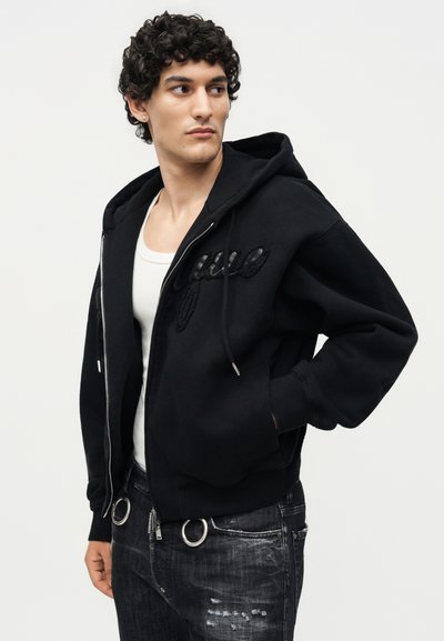 Νέος άνδρας με σγουρά μαλλιά που φοράει μαύρη ζακέτα με φερμουάρ πάνω από λευκή φανέλα και ξεθωριασμένο μαύρο τζιν, κοιτάζει προς τα πλάγια.