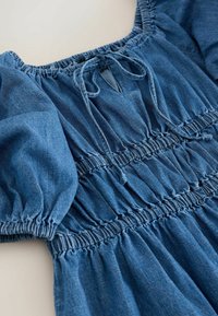 Robe en denim bleu avec manches bouffantes, taille smockée élastique et encolure, avec un détail de nouage à l'avant près du col.