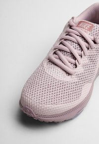 Zapatilla deportiva de Nike en material de malla rosa claro. Presenta una parte superior texturizada y cierre de cordones, con una puntera redondeada y una suela de goma.