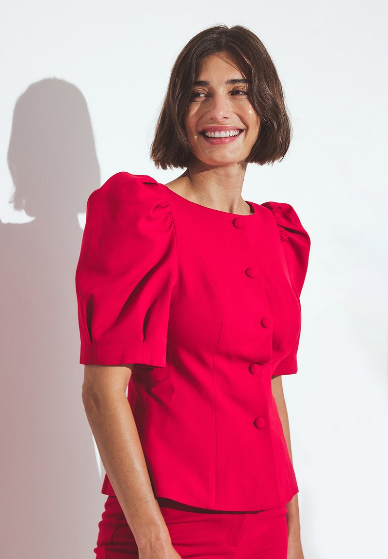 Top rosso con maniche a sbuffo, dotato di bottoni rotondi e silhouette aderente. Tessuto liscio, design strutturato e cuciture piatte. Colore uniforme senza motivi.
