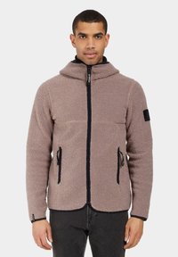 Chaqueta de forro polar en marrón claro con un cuello alto, cremallera frontal y dos bolsillos laterales. Presenta detalles en negro y una superficie texturizada.