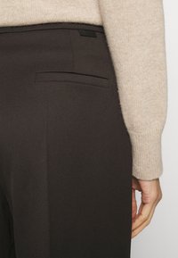 Pantalon marron sur mesure avec une finition texturée, comportant une poche arrière et une petite étiquette de logo noire. Manche de pull beige doux visible.