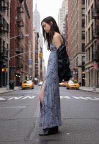 Dlouhé šedé šaty s květinovým potiskem a bočním rozparkem, doplněné černou koženou bundou. Modelka stojí na městské ulici s budovami v pozadí.