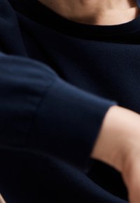 Detail navy modrého pleteného svetru s vroubkovaným manžetou a hladkou texturou, těsně přiléhající kolem paže a krku.