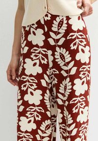 Pantalons larges en tissu bordeaux riche avec de grands motifs floraux crème et des accents de feuilles vertes, présentant une surface texturée et une tombée douce.