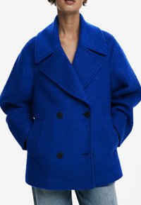 Manteau double boutonné bleu vif en tissu texturé, avec un large col, deux poches avant et des boutons noirs.