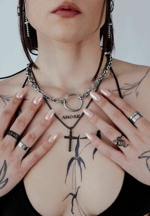 Klobige Silberketten-Halskette mit runden Anhänger, überlagert mit einem schwarzen Kreuzanhänger, ergänzt durch verschiedene Ringe und filigrane Tattoos.