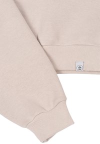 Beež huppertis, millel on soonikkoesed varrukad ja väike silt logoga. Kangas näeb välja pehme ja tekstureeritud.