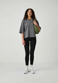 Femme aux cheveux bouclés portant un t-shirt gris surdimensionné, un legging noir, des baskets blanches, portant un sac à bandoulière vert, se tenant devant un fond uni.