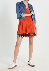 Robe orange vif avec des broderies noires à l'ourlet, associée à une veste en denim bleu clair et des baskets blanches avec des semelles marron.