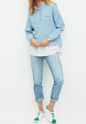 Sudadera azul clara con bolsillo frontal y capucha con cordón, combinada con una camisa blanca. Lleva unos jeans azul claros remangados y zapatillas con acentos verdes.
