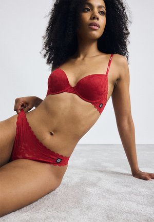 Red lace bra with curved cups, scalloped edges, and adjustable straps. Matching red lace brief with a logo tag and scalloped hem.