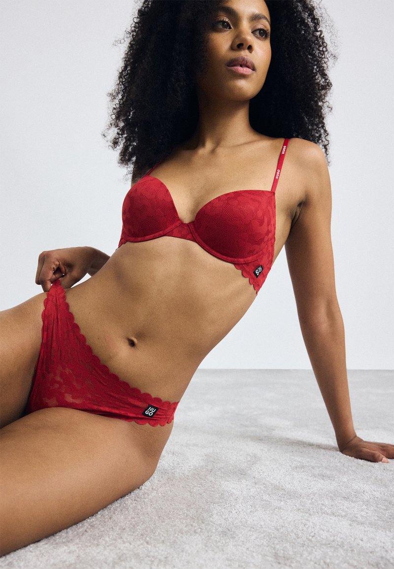 Red lace bra with curved cups, scalloped edges, and adjustable straps. Matching red lace brief with a logo tag and scalloped hem.
