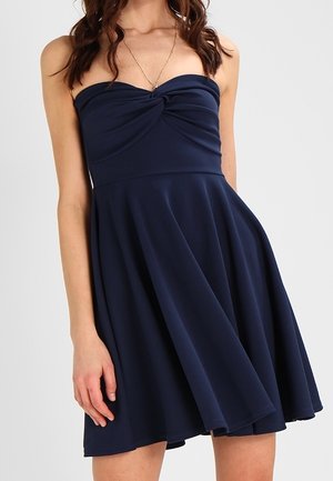 Femme portant une robe bustier bleu marine avec un nœud torsadé au niveau du buste, une jupe évasée et un collier délicat en or.