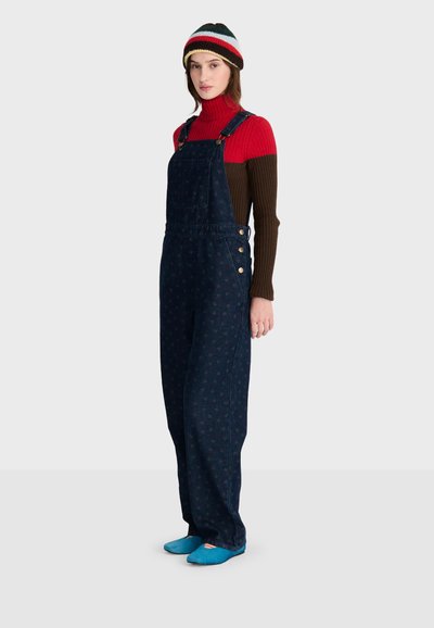 Femme portant un bonnet rayé, un pull à col roulé rouge et marron, une salopette bleu foncé à motifs, et des chaussons bleus, debout devant un fond uni.