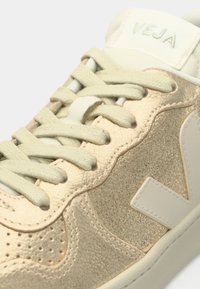 Μπεζ αθλητικό παπούτσι με υφασμένο σουέντ επάνω μέρος, ανοιχτοπράσινα κορδόνια και αντιθετικές αποχρώσεις εκρού. Διαθέτει οπές και λεπτομέρεια λογότυπου.