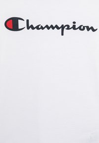 Fehér pamut póló, amelyen egy fekete, dőlt betűs "Champion" logó látható, a "C" betűben egy kis piros részlettel. Simább textúrájú és kerek nyakkivágású.