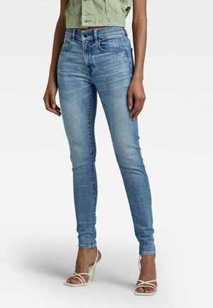 Femme portant un jean skinny bleu clair, une veste courte verte et des sandales blanches à talons hauts avec des brides, debout sur un fond blanc.