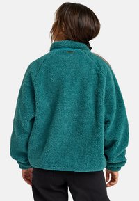 Teal fleece jas met hoge kraag, ontspannen pasvorm en gestructureerd oppervlak. Beschikt over raglanmouwen en een merklabel aan de achterkant van de nek.