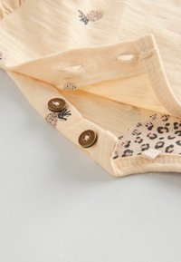 Lys ferskenfarvet stof med ananastryk, der har to brune knapper på skulderen. Indersiden har et leopardmønstret design. Blød tekstur.