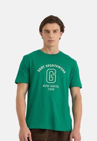 Camiseta de algodón verde de manga corta, con un gráfico blanco que dice "GANT SPORTSWEAR NEW HAVEN 1949" en la parte frontal con una tipografía en negrita.