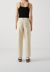 Pantalon beige clair à jambes larges, taille haute, poches arrière, texture lisse, associé à des baskets noires et blanches.