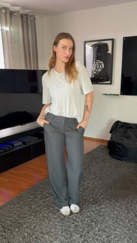 Šviesiai pilka marškinėliai, laisvai krentančios pilkos kelnės su juosmens juosta, kišenėmis ir tiesiu kirpimu. Balti sportbačiai, vidinė aplinka su tamsiais baldais.