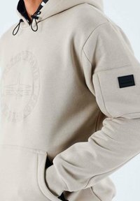 Sweatshirt beige à capuche avec un emblème texturé sur la poitrine, cordon de serrage ajustable et un patch noir sur la manche. Tissu doux.