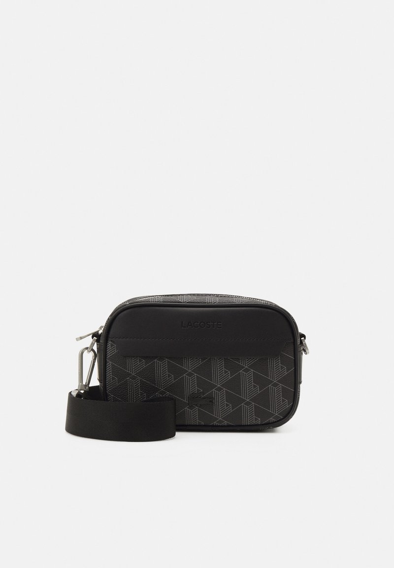 Piccola borsa a tracolla nera con accessori in argento, motivo geometrico, logo "Lacoste" in rilievo e tracolla regolabile su sfondo bianco.