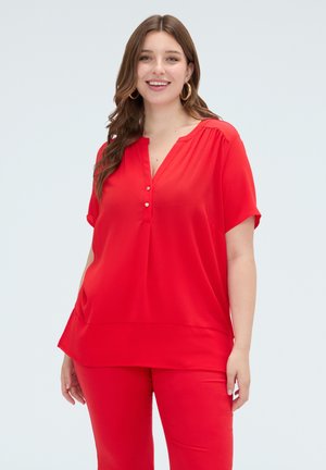 Vrouw met lang bruin haar, gouden oorringen in hoepelvorm, een rood blouse met korte mouwen en knopen en een rode broek, glimlachend en staand.