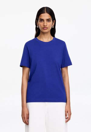 Donna con lunghi capelli scuri indossa una maglietta semplice royal blue a maniche corte e pantaloni bianchi, rivolta verso l'obiettivo su sfondo bianco.