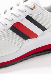 Chaussure de sport blanche avec un tissu en maille, lacets blancs et détails rayés noir, blanc et rouge sur le côté, ainsi qu'une semelle rayée rouge.