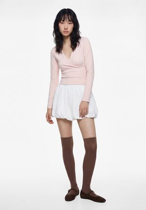 Jonge vrouw met lang zwart haar, gekleed in een lichtroze wikkeltrui met lange mouwen, een witte bubbelsrok, bruine kniehoge sokken en bruine schoenen.