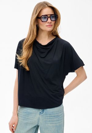 Kobieta z długimi blond włosami, nosząca oversize'owe okulary przeciwsłoneczne w oprawkach typu żółw, luźną czarną bluzkę z krótkim rękawem oraz jasnoniebieskie dżinsy, pozująca z ręką na biodrze.