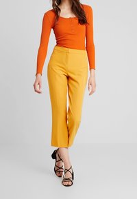 Top de manga larga acanalado naranja con detalles de botones, combinado con pantalones cortos amarillos de talle alto y tacones negros con tiras.