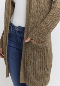 Bežs adīts kardigāns ar teksturētu rakstu, atvērta priekšpuse un sānu kabatas, kombinēts ar ziliem džinsu biksēm.