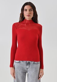 Rotes geripptes Rollkragensweatshirt mit durchsichtigen horizontalen Streifen am Ausschnitt, kombiniert mit grauen Jeans.