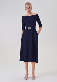 Marineblaues Off-Shoulder-Kleid mit Dreiviertelärmeln, tailliertem Schnitt und dekorativem goldenen Schnallenakzent. Glatter Stoff mit einem ausgestellten Rock.