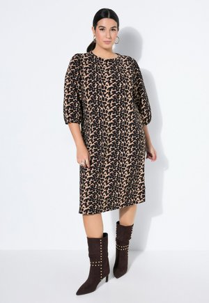 Kvinde stående mod hvid baggrund iført en knælængde leopardprint kjole med ballonærmer og sorte nittede hæle støvler.